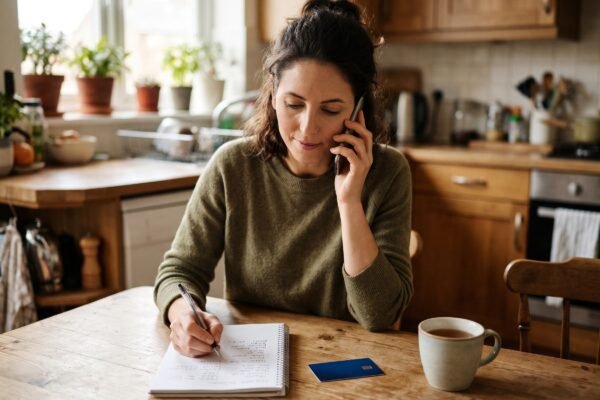 A woman sitting at her kitchen table on the phone with a credit card and notepad, negotiating a lower interest rate with her card issuer
